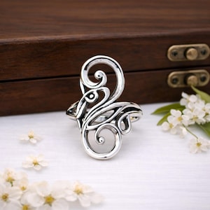 May include: A silver-colored ring with an ornate, swirling design. The ring features a central, stylized shape with curved lines and circular accents. The ring is set against a white surface with small white flowers and a wooden box in the background.
