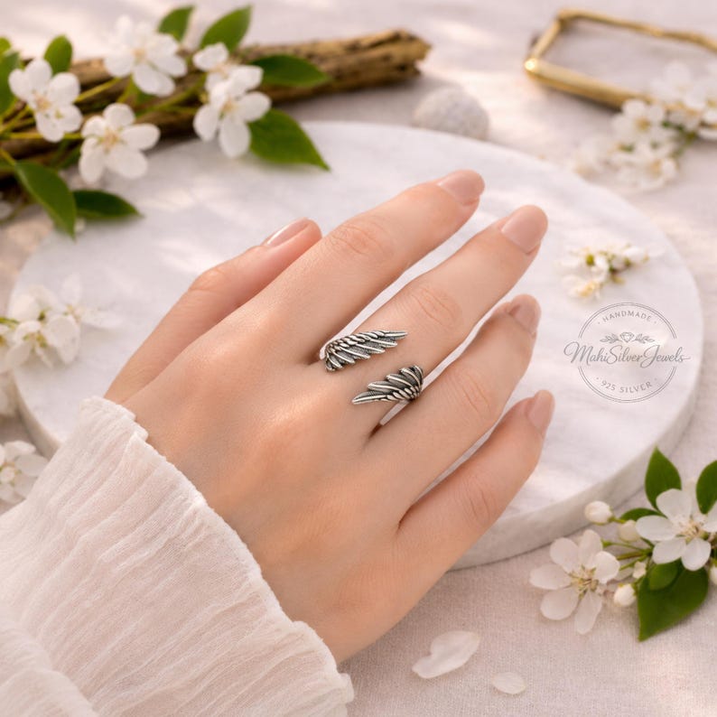 K&ouml;nnte beinhalten: Ein offener Silberring mit zwei detaillierten, gefiederten Fl&uuml;gelmotiven, die einander zugewandt sind. Das Metall hat ein oxidiertes Finish, um die Textur der einzelnen Federn hervorzuheben. Der Ring wird am Finger getragen und zeigt das Design.
