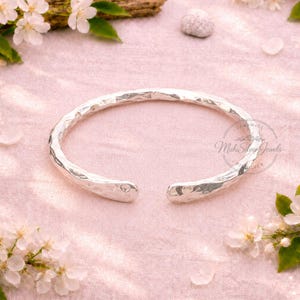 May include: A hammered silver cuff bracelet with an open design. The bracelet has a textured surface and is displayed on a pink surface with white flowers and petals. The text "Mahi Silver Jewels" is visible.