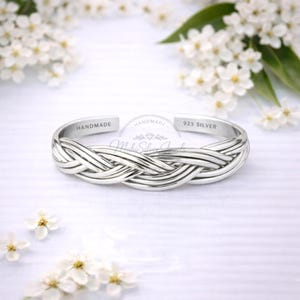 Puede incluir: Una pulsera de plata con diseño trenzado. La pulsera está estampada con "HANDMADE" y "925 SILVER". La joya se exhibe sobre una superficie blanca con flores blancas en el fondo. Un accesorio elegante.
