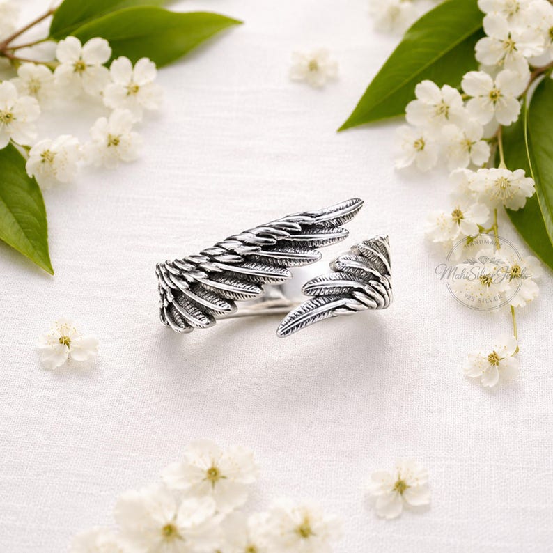 K&ouml;nnte beinhalten: Ein silberner Ring in Form von zwei &uuml;berlappenden Engelsfl&uuml;geln mit detaillierter Federstruktur und oxidierter Oberfl&auml;che. Der Metallring ist offen gestaltet und l&auml;sst sich anpassen. Ein kreisf&ouml;rmiger Logostempel mit dem Text Mahi Silver Jewels 925 ist sichtbar.