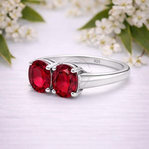 Op de afbeelding: Een zilveren ring met twee ovaal geslepen, helderrode edelstenen. De ring is geplaatst op een witte ondergrond met vage witte bloemen en groene bladeren op de achtergrond. De band is gepolijst, met een 925-stempel.