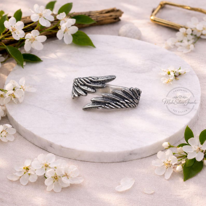 K&ouml;nnte beinhalten: Ein Silberring mit zwei detaillierten, &uuml;berlappenden Fl&uuml;gelmotiven und oxidierten Akzenten, die die Federstruktur hervorheben. Der Ring ist mit dem Text Mahi Silver Jewels 925 Silver gepr&auml;gt, was auf seine Zusammensetzung aus Sterlingsilber hinweist.