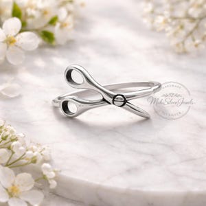 May include: A silver-colored ring shaped like a pair of scissors. The ring has a scissor design with two loops and blades. The ring is on a white marble surface with white flowers in the background. The text "HANDMADE Mahi Silver Jewels 925 SILVER" is visible.