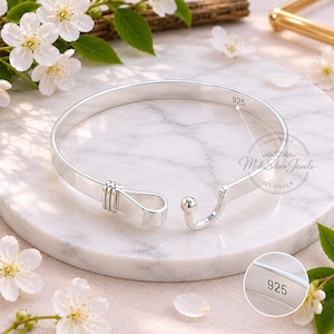 May include: A silver bangle bracelet with a hook and eye closure. The bracelet is marked with "925" and "Mahi Silver Jewels". The open design features a rounded band with a hook closure on one end and a small ball on the other. The bracelet is displayed on a marble surface.