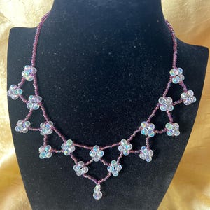 Puede incluir: Un collar de cuentas con un diseño floral. El collar presenta cuentas claras e iridiscentes dispuestas en forma de flor, conectadas por una cadena de cuentas moradas. Se exhibe en un maniquí negro.