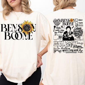 Könnte beinhalten: Elfenbeinfarbenes T-Shirt mit dem Text "BENSON BOONE" und einer Sonnenblumen-Grafik. Auf der Rückseite des Shirts befinden sich zusätzliche Grafiken und Texte, darunter "Talk with me for Days" und "darling you're my work of art."