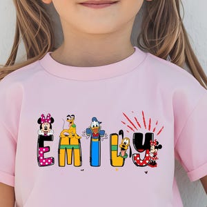 Op de afbeelding: Een lichtroze T-shirt met de naam "EMILY" in kleurrijke, cartoonachtige letters. Elke letter is versierd met Disney-personages, waaronder Minnie Mouse, Goofy, Donald Duck en Mickey Mouse. De "U" heeft rode accenten.