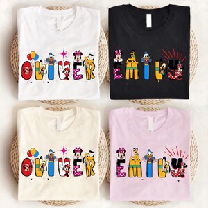 Op de afbeelding: Vier T-shirts, wit, zwart, cr&egrave;me en roze, elk met een andere naam in kleurrijke letters met Disney-thema. De namen zijn "Oliver" en "Emily", met stripfiguren ge&iuml;ntegreerd in de letters. De shirts zijn op gevlochten matten gevouwen.