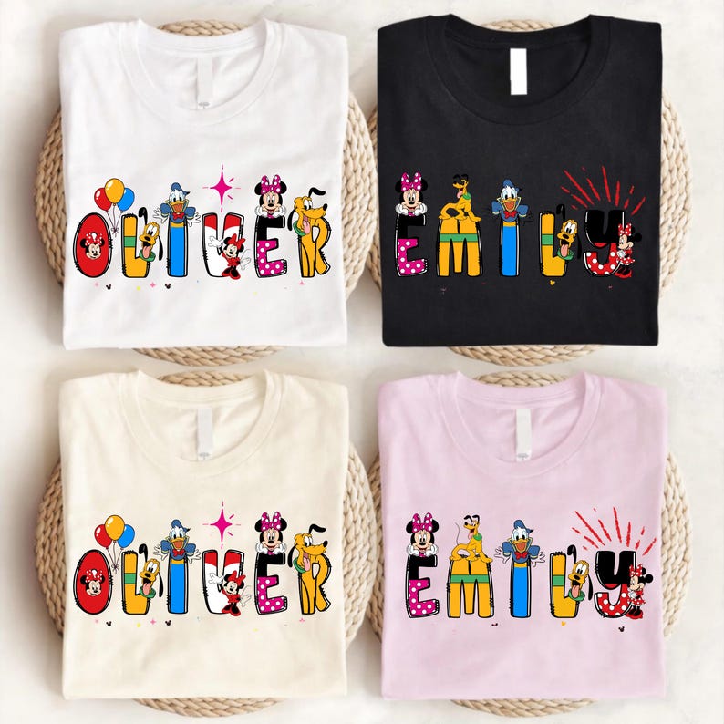 Op de afbeelding: Vier T-shirts, wit, zwart, cr&egrave;me en roze, elk met een andere naam in kleurrijke letters met Disney-thema. De namen zijn "Oliver" en "Emily", met stripfiguren ge&iuml;ntegreerd in de letters. De shirts zijn op gevlochten matten gevouwen.
