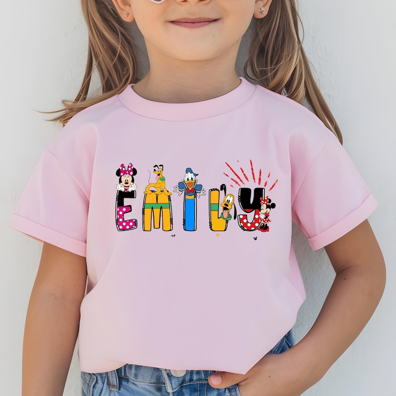 Op de afbeelding: Een lichtroze T-shirt met de naam "EMILY" in kleurrijke, cartoonachtige letters. Elke letter is versierd met Disney-personages, waaronder Minnie Mouse, Goofy, Donald Duck en Mickey Mouse. De "U" heeft rode accenten.