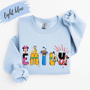 Op de afbeelding: Lichtblauw sweatshirt met de naam "EMILY" in kleurrijke letters met Disney-thema. De letters bevatten Minnie Mouse, Pluto, Donald Duck en Goofy. De woorden "light blue" zijn cursief geschreven in de linkerbovenhoek.