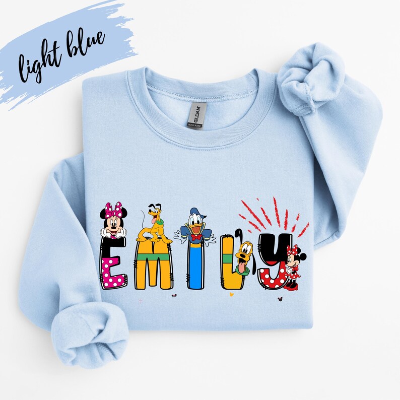 Op de afbeelding: Lichtblauw sweatshirt met de naam "EMILY" in kleurrijke letters met Disney-thema. De letters bevatten Minnie Mouse, Pluto, Donald Duck en Goofy. De woorden "light blue" zijn cursief geschreven in de linkerbovenhoek.