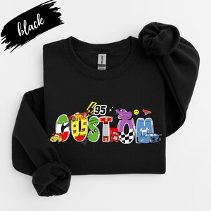Op de afbeelding: Zwart sweatshirt met het woord "CUSTOM" in kleurrijke, cartoonachtige letters, met auto-illustraties. Het nummer "95" staat boven het woord. Het woord "black" is in cursiefschrift linksboven geschreven.