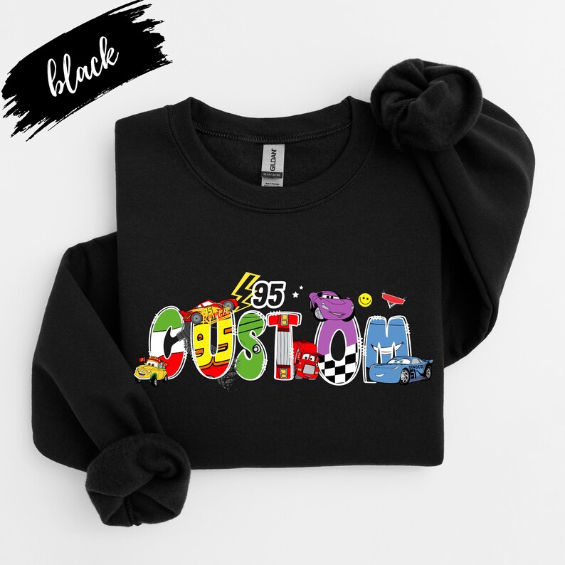Op de afbeelding: Zwart sweatshirt met het woord "CUSTOM" in kleurrijke, cartoonachtige letters, met auto-illustraties. Het nummer "95" staat boven het woord. Het woord "black" is in cursiefschrift linksboven geschreven.