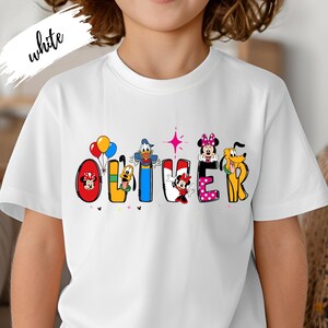 Op de afbeelding: Wit T-shirt met de naam "OLIVER" in kleurrijke Disney-letters. Elke letter is versierd met een ander Disney-personage, waaronder Mickey en Minnie Mouse, Donald Duck en Pluto. Het woord "white" is in cursiefschrift linksboven geschreven.