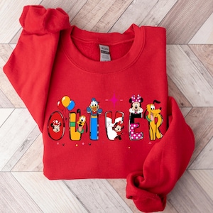 Op de afbeelding: Een rode sweatshirt met de naam "OLIVER" in kleurrijke Disney-letters. Elke letter bevat een ander Disney-personage, waaronder Mickey Mouse, Donald Duck en Minnie Mouse. De sweatshirt is gemaakt van een zacht materiaal.