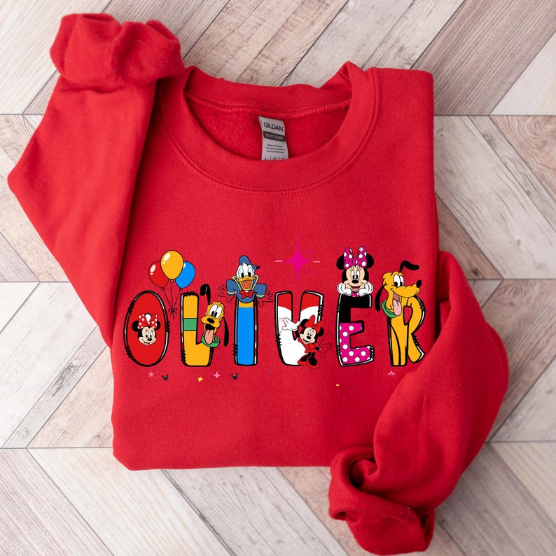 Op de afbeelding: Een rode sweatshirt met de naam "OLIVER" in kleurrijke Disney-letters. Elke letter bevat een ander Disney-personage, waaronder Mickey Mouse, Donald Duck en Minnie Mouse. De sweatshirt is gemaakt van een zacht materiaal.