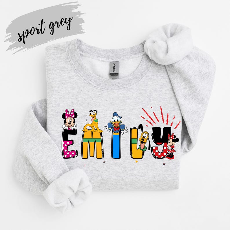 Op de afbeelding: Een lichtgrijs sweatshirt met de naam "EMILY" in kleurrijke letters met Disney-thema. Minnie Mouse, Donald Duck en Pluto zijn afgebeeld. De woorden "sport grey" staan in cursiefschrift linksboven.