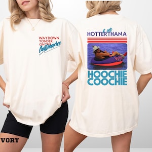 May include: Cream-coloured t-shirt with text on the front and back. The front says "WAYDOWN YONDER ON THE Chattahoochee" in red and blue. The back features a photo of a person in a tube with the text "HOTTER THANA HOOCHIE COOCHIE".