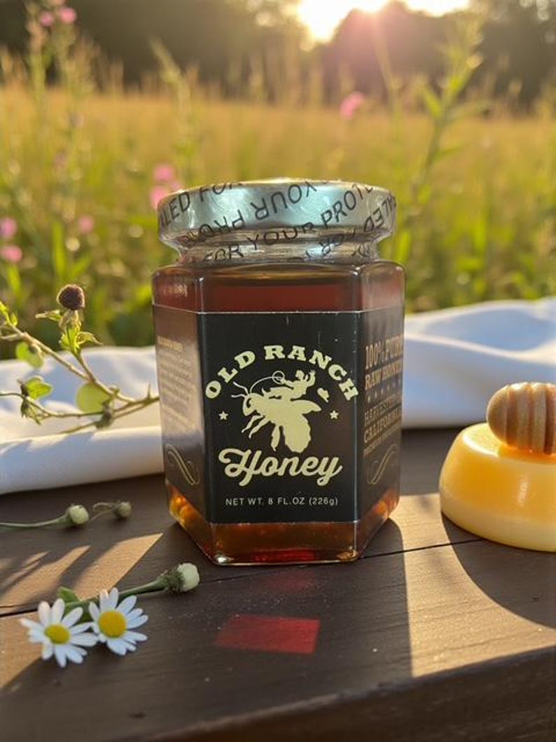 Puede incluir: Un tarro hexagonal de vidrio de miel Old Ranch, con tapa y etiqueta doradas. La etiqueta presenta una ilustraci&oacute;n de abeja y el texto "Honey". El tarro contiene 226g de miel. La imagen est&aacute; al aire libre con flores y sol.
