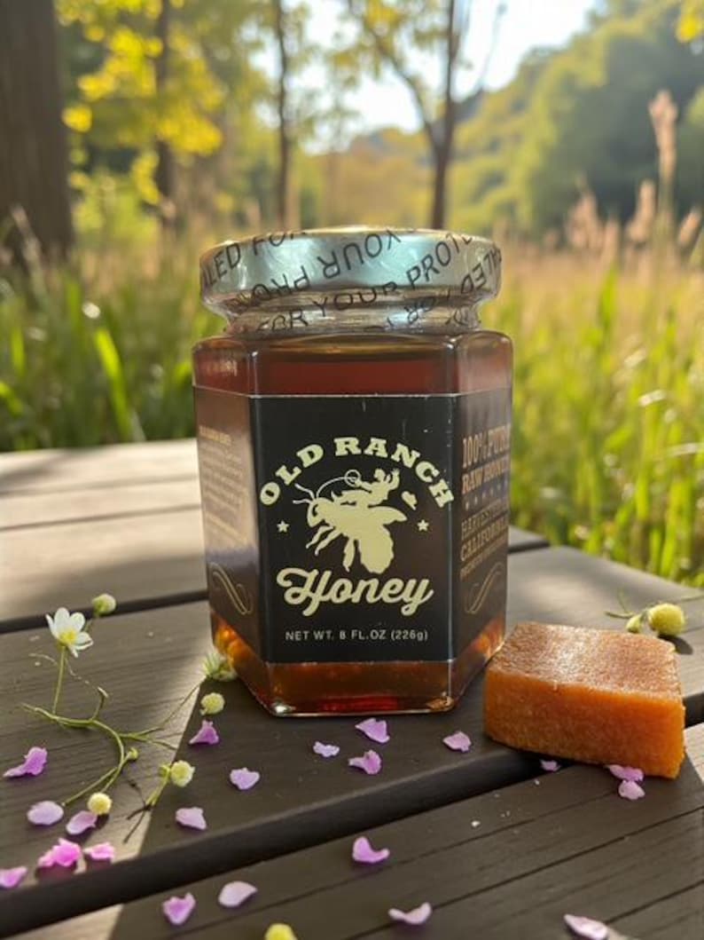 Puede incluir: Un tarro de miel Old Ranch, 226g, con un gr&aacute;fico de abeja. La miel es de color &aacute;mbar y se encuentra sobre una superficie de madera con un trozo de comida naranja-marr&oacute;n. El fondo est&aacute; borroso con follaje verde.