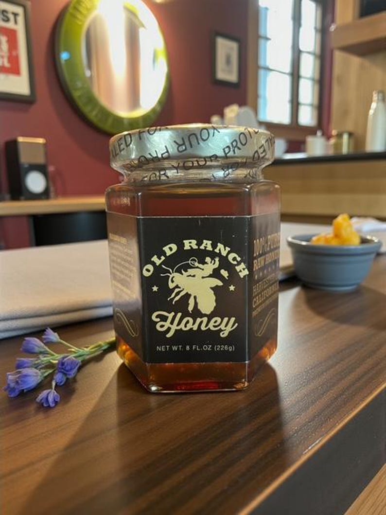 Puede incluir: Un tarro hexagonal de vidrio de miel Old Ranch, que contiene miel de color &aacute;mbar. La etiqueta presenta una ilustraci&oacute;n de abeja y el texto "Honey, Net Wt. 8 fl oz (226g)". El tarro est&aacute; sellado y sobre una superficie de madera.