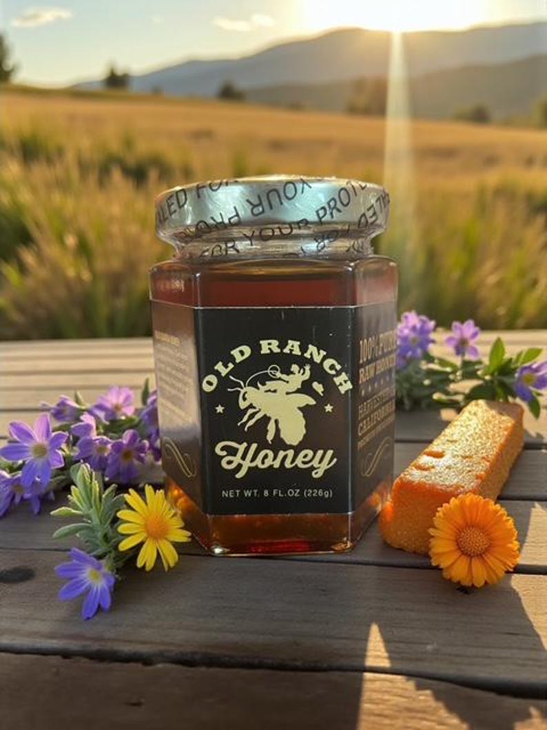 Puede incluir: Un tarro de miel Old Ranch, con un logotipo de abeja, se muestra. La etiqueta dice "100% Pure Raw Honey". El tarro est&aacute; junto a un trozo de pastel de naranja y flores. El peso neto es de 226 g.