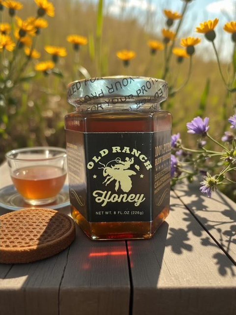 Puede incluir: Un tarro de miel Old Ranch, con un gr&aacute;fico de abeja, est&aacute; sobre una superficie de madera. El tarro contiene 226g de miel. Una taza de t&eacute; y una galleta est&aacute;n al lado del tarro. Flores silvestres amarillas y azules en el fondo.