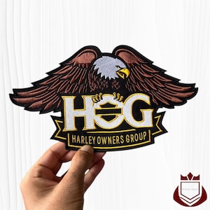May include: Embroidered patch featuring a brown and black eagle with outstretched wings, clutching the letters "HOG" in white and gold. The text "HARLEY OWNERS GROUP" is below. The patch is held by a hand.
