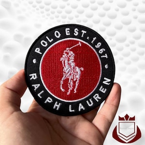 May include: Round black and red embroidered patch with the text "POLO EST. 1967 RALPH LAUREN" surrounding a white polo player logo. The patch is held in a hand.