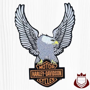 May include: Embroidered patch featuring a majestic eagle with outstretched wings. The eagle is gray and white, with a yellow beak and talons. Below the eagle is a black and orange Harley-Davidson logo with the words "MOTOR HARLEY-DAVIDSON CYCLES".