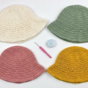 May include: Four crocheted bucket hats in cream, sage green, dusty rose, and mustard yellow. The arrangement includes four matching skeins of yarn, two pink crochet hooks, a small circular measuring tape, and a pair of pink-handled scissors.