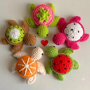 May include: Five crocheted turtle plushies shaped like fruits, including a kiwi, strawberry, dragon fruit, orange, and watermelon, arranged around a small red heart. Each turtle features a textured shell pattern and a soft, yarn-based construction.