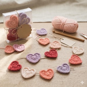 Peut inclure: Une boîte en plastique transparent contenant plusieurs cœurs crochetés dans des tons de rose, rouge et violet, attachés avec de la ficelle et une étiquette vierge. Une guirlande de cœurs crochetés assortis, une pelote de laine rose et un crochet en bois.