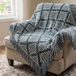 Puede incluir: Una manta de ganchillo con un patrón geométrico de cuadrícula en tonos azul y crema. La manta tiene una superficie texturizada con puntos en relieve que forman rombos y está rematada con flecos largos y gruesos en los bordes.