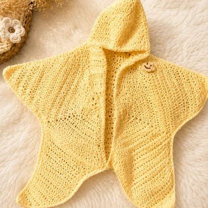 May include: A pale yellow star-shaped hooded blanket made of crocheted yarn. The garment features a pointed hood and a small, circular wooden button with a carved smiley face detail attached to the upper right side of the front opening.
