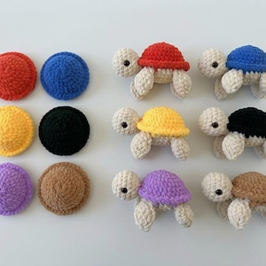 May include: A collection of six crocheted turtle figurines and six matching circular crocheted shells in red, blue, yellow, black, purple, and brown. Each turtle features a cream-colored head and limbs with small black eyes and a colorful shell.