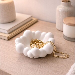 Peut inclure: Un vide-poches blanc en forme de nuage contient un collier en chaîne dorée. Le plat est posé sur une surface en bois clair, à côté d'une lampe, de livres et d'un vase décoratif. Le vide-poches est un objet décoratif pour ranger des bijoux.