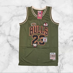 Op de afbeelding: Olijfgroen basketbalshirt met het woord "BULLS" en het nummer "23" in oranje en zwart. Het shirt heeft een crèmekleurige halslijn en armsgaten, en diverse patches. Een label is bovenaan bevestigd.