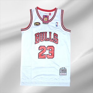 Op de afbeelding: Wit basketbalshirt met rode en zwarte accenten. Het shirt toont het woord "BULLS" en het nummer "23" in rood. Inclusief NBA Finals en Mitchell & Ness labels. Een klassiek sportkledingstuk.