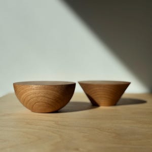 May include: Two small, wooden bowls with a natural finish sit on a light-colored wooden surface. One bowl is a half-sphere shape, and the other is a truncated cone. The bowls are made of a light-colored wood.