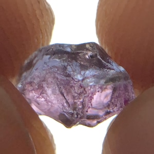 May include: A raw, translucent purple gemstone with an irregular, multifaceted shape. The stone displays varying shades of violet and lavender, with internal inclusions and a natural, unpolished texture that catches the light from behind.
