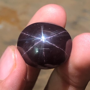 Può includere: Una gemma cabochon lucida di colore viola scuro intenso che presenta un distinto effetto a stella a sei raggi, noto come asterismo. La superficie liscia e arrotondata della pietra mostra un riflesso luminoso centrato a forma di stella.
