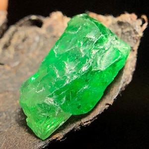 May include: A raw, vibrant green gemstone with a jagged, crystalline structure and translucent facets. The stone displays a deep emerald hue with natural surface irregularities and sharp, angular edges, reflecting light across its uneven, textured surface.
