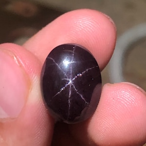 Può includere: Una gemma cabochon di forma ovale color viola scuro, caratterizzata da un distinto motivo a stella a sei punte sulla sua superficie liscia e lucida. La pietra presenta un colore profondo e saturo con un effetto di luce riflettente.