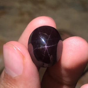 Può includere: Una gemma cabochon di forma ovale, di colore viola scuro intenso, caratterizzata da un distinto motivo a stella a sei punte sulla sua superficie liscia e lucida. La pietra presenta una lucentezza riflettente e un effetto stellato.