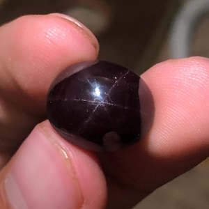 Può includere: Una gemma cabochon di forma ovale, di colore viola scuro e profondo, caratterizzata da un distinto motivo a stella a sei punte di colore chiaro sulla sua superficie liscia e lucida. La pietra presenta una lucentezza riflettente.