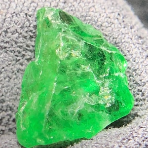 May include: A raw, unpolished gemstone with a vibrant, translucent emerald green color. The stone features an irregular, jagged shape with multiple facets and natural internal inclusions that catch the light, creating a bright, crystalline appearance.