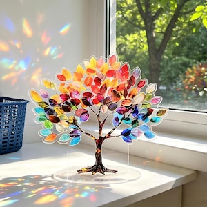 Op de afbeelding: Een decoratieve boomsculptuur met kleurrijke bladeren in tinten rood, oranje, geel, groen, blauw en paars. De boom heeft een bruine stam en takken en staat op een heldere, ronde basis. De bladeren zijn doorschijnend.